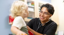 A smiling clinician uses a stethoscope to examine a young toddler in a pediatric exam room.