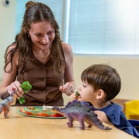 Adult smiles while encouraging a young child to eat vegetables at a table, using toy dinosaurs during a snack activity.