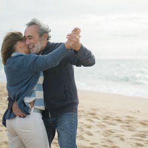 An older couple smiles and dances together on a sandy beach, holding each other close with ocean waves in the background.