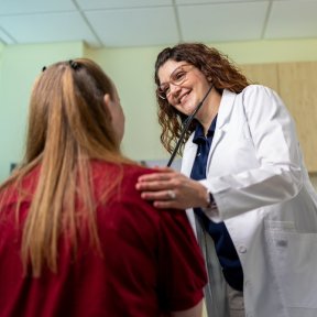 Doctor in a white coat smiles while listening to a patient’s chest with a stethoscope during an exam in a clinic room.