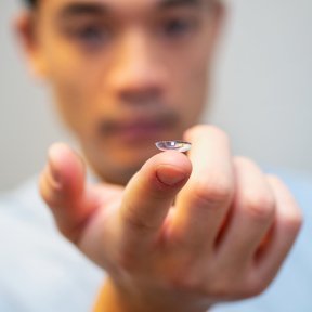 A close-up of a contact lens balanced on a student's fingertip, with the student's face softly blurred in the background.