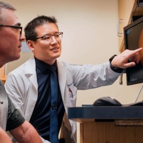 An optometrist in a white coat points to a retinal scan image on a monitor while reviewing results with a patient.