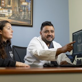 A dental professional in a white coat points to imaging on a monitor while a patient looks on in a clinical office.