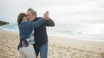 An older couple smiles and dances together on a sandy beach, holding each other close with ocean waves in the background.