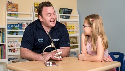 Speech-language pathologist uses a mouth model and mirror while working with a young girl.