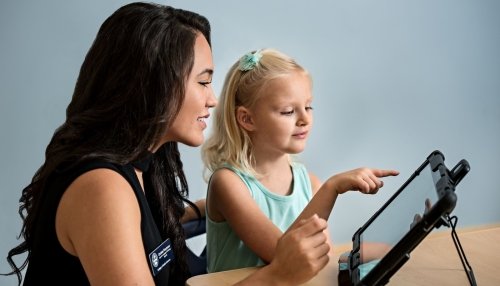 Young girl points to tablet screen while working with a Speech-Language Pathologist.