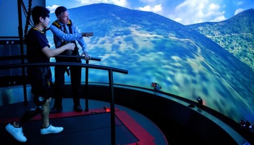 Man in harness walking in front of CAREN virtual screen showing mountains. A doctor is standing to the side and pointing. 