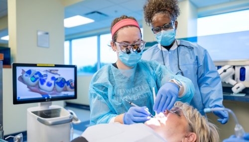 Dental students wearing protective gear perform a procedure on a patient, with a digital tooth scan displayed on a nearby monitor.