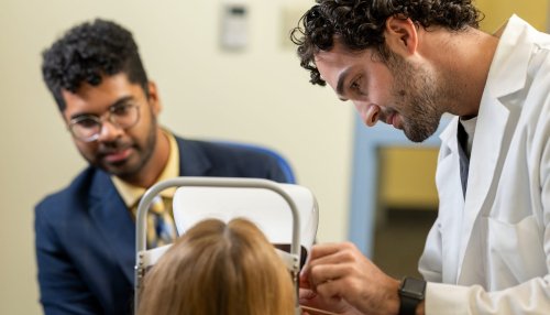 An optometry student in a white coat performs a slit-lamp eye examination on a patient while an instructor observes.