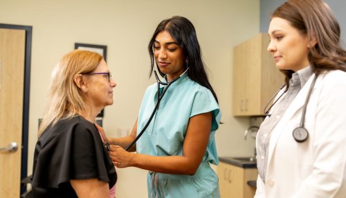 A medical student in scrubs uses a stethoscope on a patient while a white-coated clinician observes in an exam room.