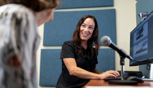 A speech-language pathologist smiles at a patient across a desk with a microphone and monitor in a sound-treated clinical room.