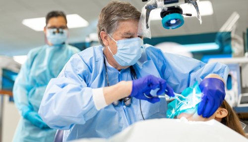 A dentist in a mask, gown, and gloves performs a dental procedure on a patient under a surgical light in a clinical setting.